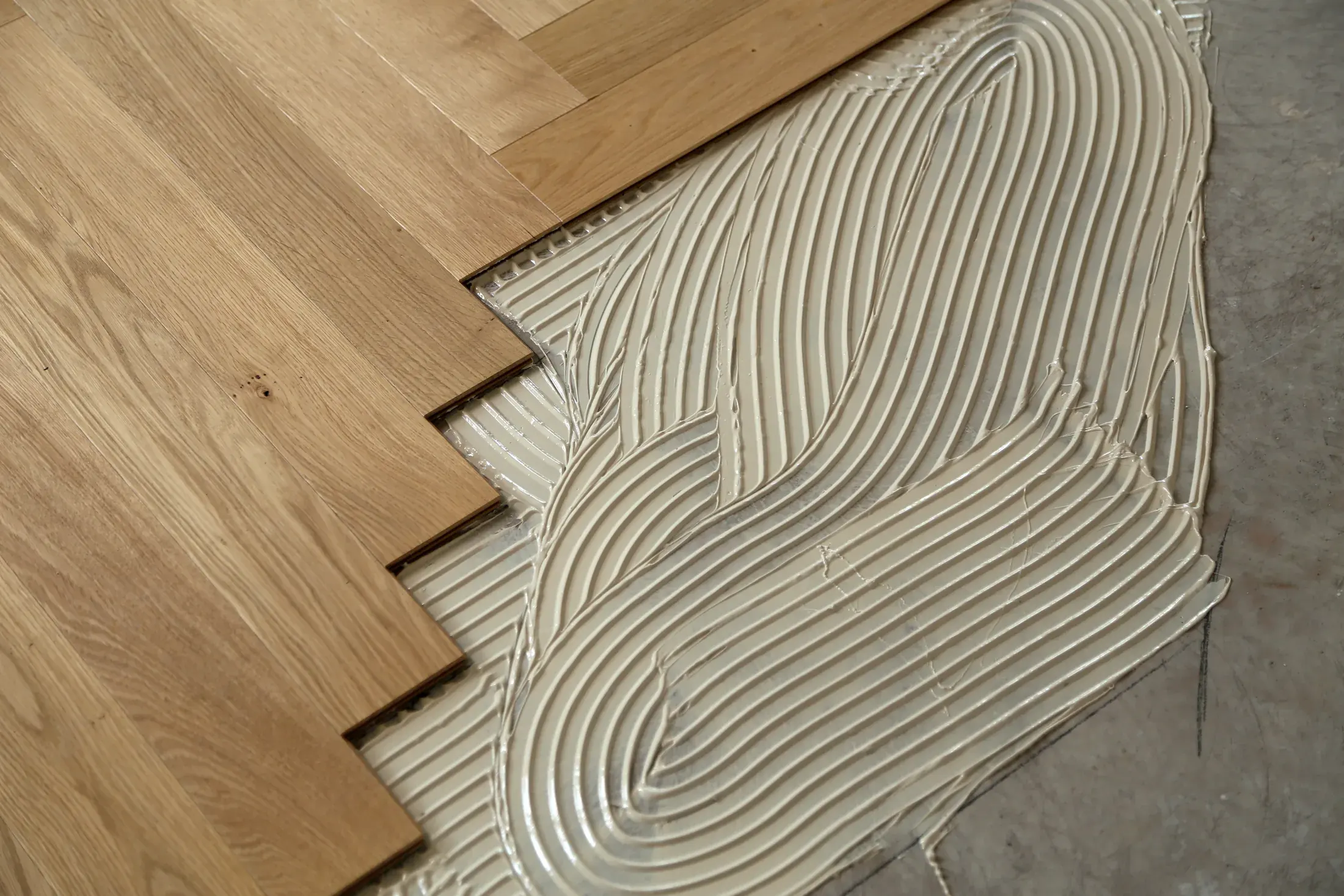 Herringbone engineered hardwood planks being laid over troweled adhesive on a concrete slab