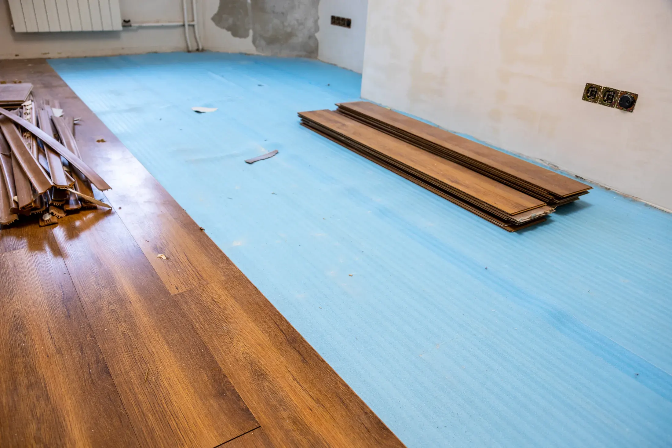 Laminate planks stacked on blue underlayment in an empty room mid-installation